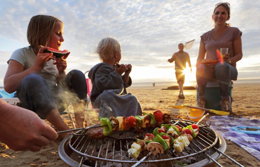 A family is enjoying grilling kebabs together on the beach as the sun begins to set.