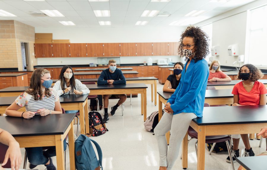 Masked high school teacher addressing students in classroom